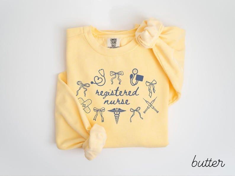 May include: A light yellow sweatshirt with the words "registered nurse" surrounded by medical-themed illustrations in navy blue. The sweatshirt is knotted at the bottom and has a matching bow on the shoulder.