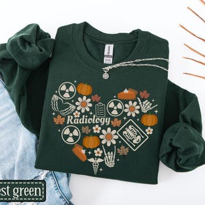 May include: A forest green crewneck sweatshirt featuring a heart-shaped graphic composed of pumpkins, autumn leaves, daisies, skeleton hands, radiation symbols, a clipboard, and a medical caduceus with the word Radiology printed in the center.