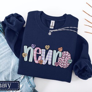 May include: Navy blue sweatshirt with the word "neur" in a colorful, playful font, and a brain graphic. The word "neur" is decorated with heart-shaped candies. A silver necklace is draped across the sweatshirt.