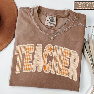 May include: A brown short-sleeved t-shirt with the word TEACHER printed in a mix of orange and white gingham, striped, and pumpkin-patterned block letters. The shirt features a Comfort Colors label at the neck and a silver-toned pendant necklace.