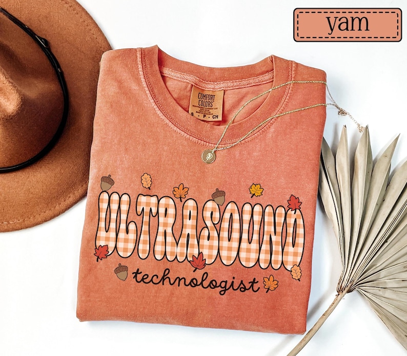 Puede incluir: Una camiseta color &ntilde;ame con las palabras "ULTRASOUND technologist" en un dise&ntilde;o a cuadros. La camiseta presenta gr&aacute;ficos con tem&aacute;tica oto&ntilde;al de bellotas y hojas. Tambi&eacute;n se ven un collar dorado y un sombrero marr&oacute;n.