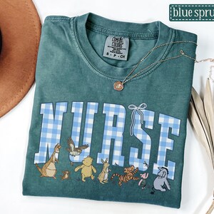 K&ouml;nnte beinhalten: Ein teal Comfort Colors T-Shirt mit dem Wort "NURSE" in gro&szlig;en, hellblau-karierten Buchstaben. Unter dem Wort befinden sich Zeichentrickfiguren. Eine goldene Halskette ist auf dem Shirt drapiert. Das Shirt ist mit "blue spruce" beschriftet.