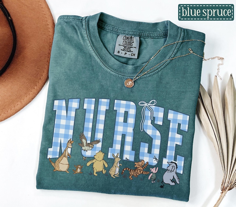K&ouml;nnte beinhalten: Ein teal Comfort Colors T-Shirt mit dem Wort "NURSE" in gro&szlig;en, hellblau-karierten Buchstaben. Unter dem Wort befinden sich Zeichentrickfiguren. Eine goldene Halskette ist auf dem Shirt drapiert. Das Shirt ist mit "blue spruce" beschriftet.