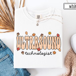 Puede incluir: Sudadera blanca de cuello redondo con las palabras "ULTRASOUND technologist" en una fuente naranja a cuadros, decorada con hojas de oto&ntilde;o y bellotas. La sudadera es de la marca Comfort Colors. La palabra "white" est&aacute; en un cuadro en la esquina superior derecha.