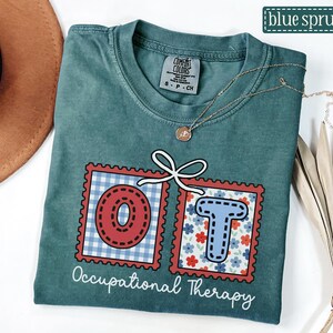 May include: Blue spruce t-shirt with the letters "OT" in red and blue postage stamp designs. The shirt has the words "Occupational Therapy" in white script. A gold necklace is draped across the shirt.