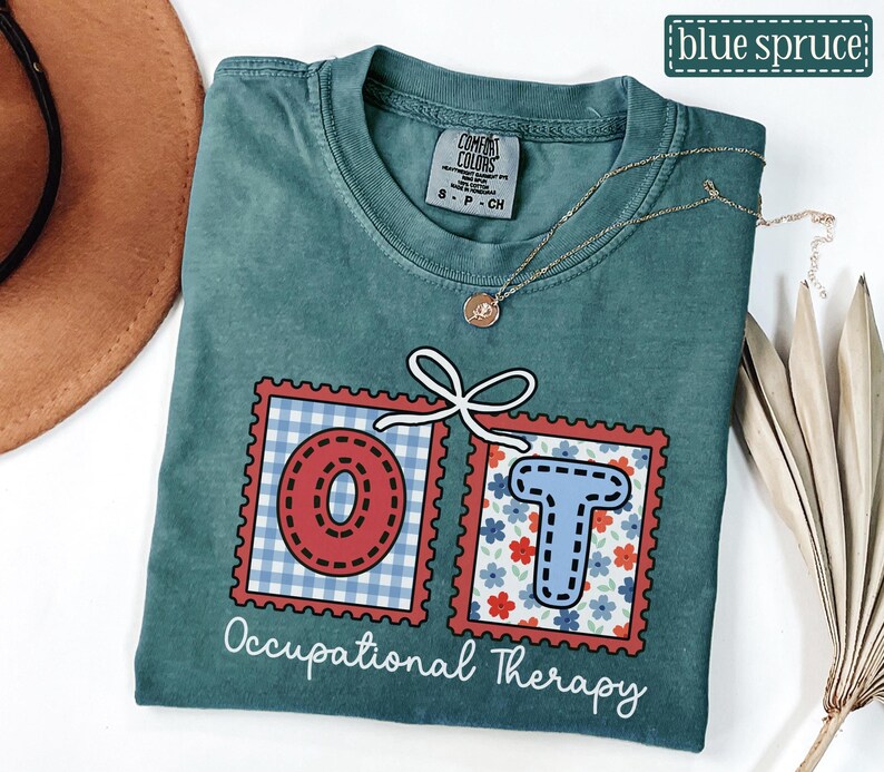 May include: Blue spruce t-shirt with the letters "OT" in red and blue postage stamp designs. The shirt has the words "Occupational Therapy" in white script. A gold necklace is draped across the shirt.