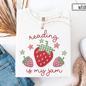May include: A white t-shirt featuring a graphic of three strawberries with green and red gingham patterns. The text "reading is my jam" is printed in red script above and below the strawberries, accompanied by small gingham star icons.