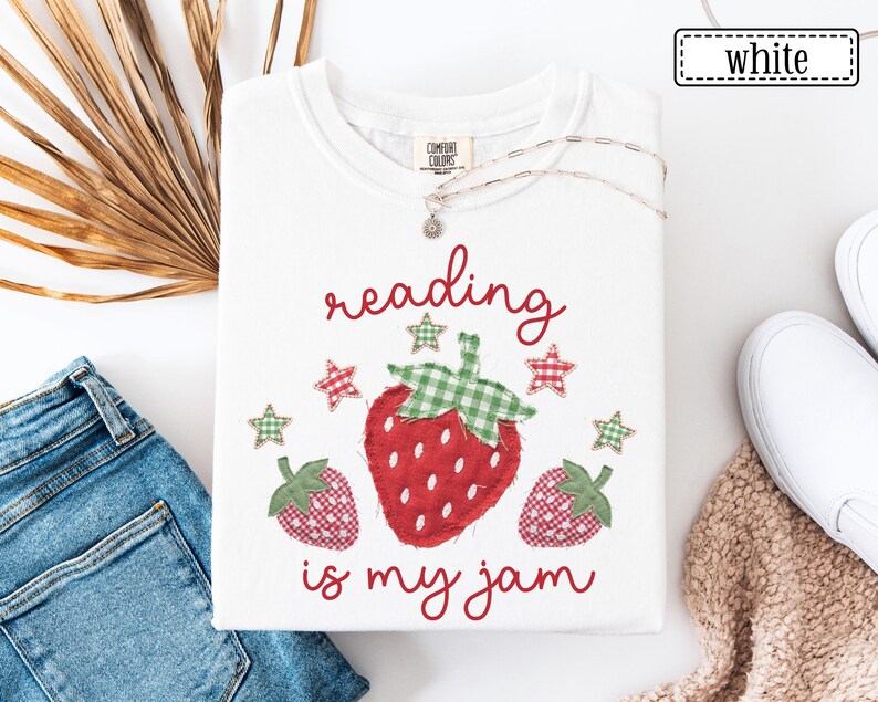 May include: A white t-shirt featuring a graphic of three strawberries with green and red gingham patterns. The text "reading is my jam" is printed in red script above and below the strawberries, accompanied by small gingham star icons.