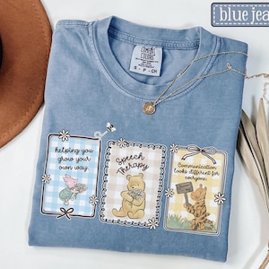 May include: Blue jean colored t-shirt with three framed illustrations featuring Winnie the Pooh characters. The frames have text that reads "helping you grow your own way", "Speech Therapy", and "Communication looks different for everyone."