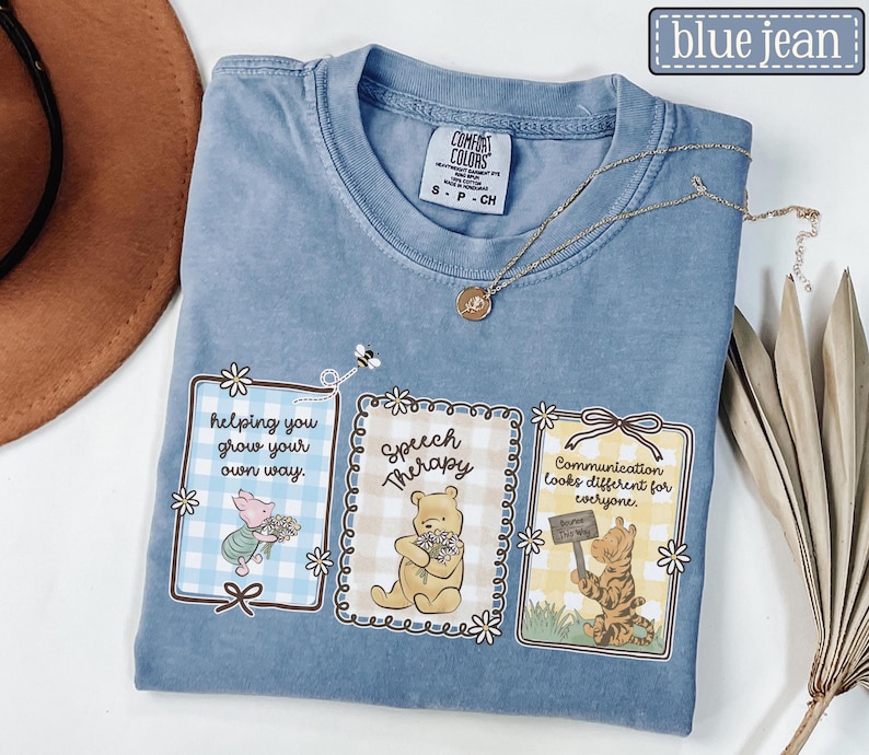 May include: Blue jean colored t-shirt with three framed illustrations featuring Winnie the Pooh characters. The frames have text that reads "helping you grow your own way", "Speech Therapy", and "Communication looks different for everyone."
