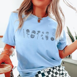 May include: A light blue t-shirt with a black line drawing of a crochet hook, yarn, flowers, and buttons. The shirt has a crew neck and short sleeves.
