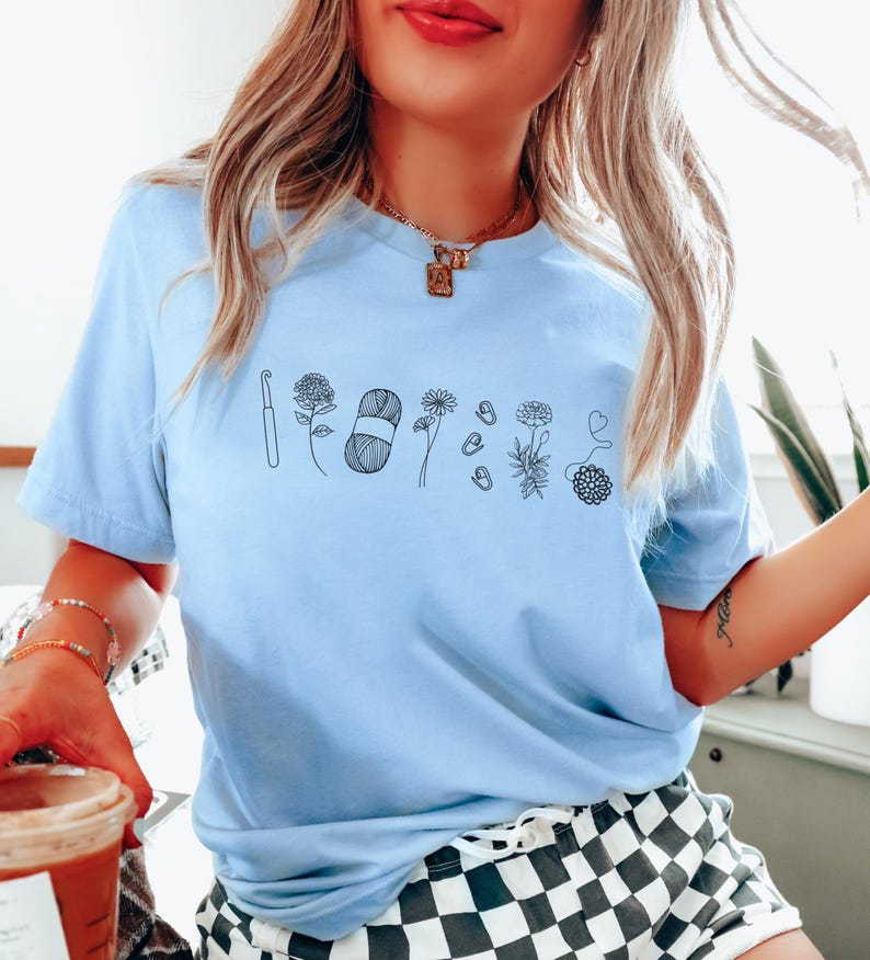 May include: A light blue t-shirt with a black line drawing of a crochet hook, yarn, flowers, and buttons. The shirt has a crew neck and short sleeves.