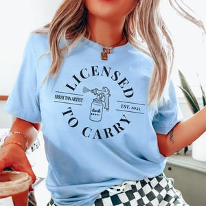 May include: A light blue t-shirt with a black circle graphic that says "Licensed to Carry" with a spray tan artist logo and the text "Spray Tan Artist" and "Est. 2025".