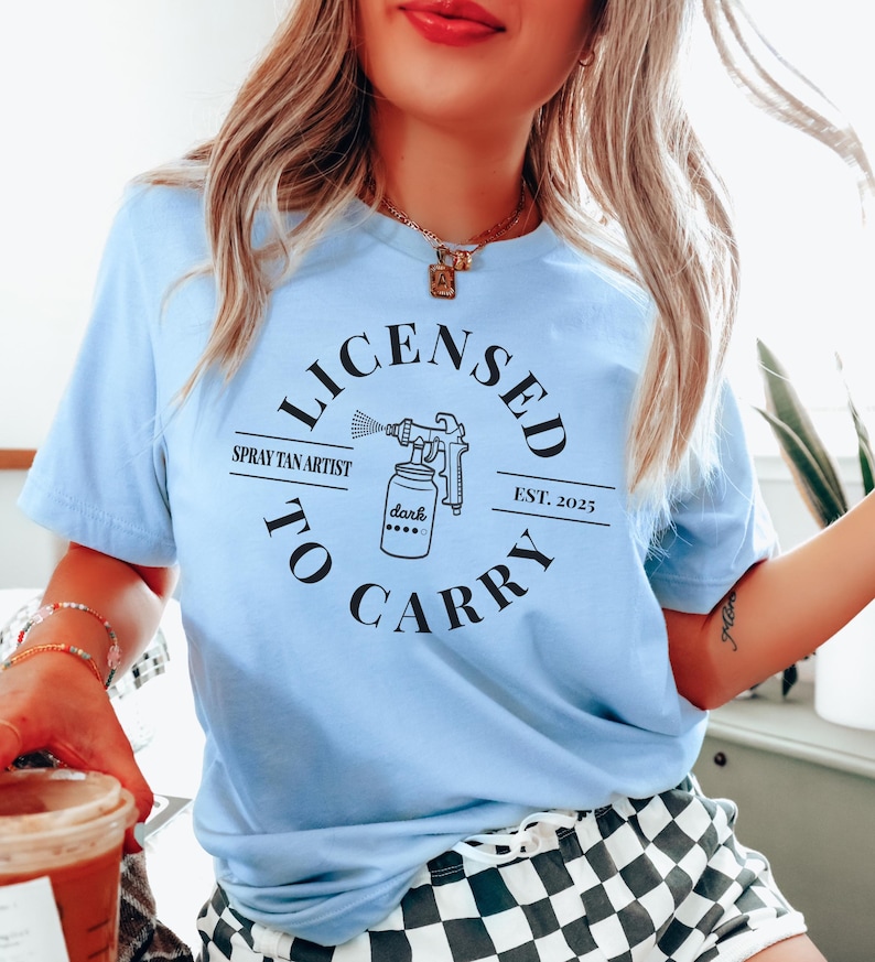 May include: A light blue t-shirt with a black circle graphic that says "Licensed to Carry" with a spray tan artist logo and the text "Spray Tan Artist" and "Est. 2025".
