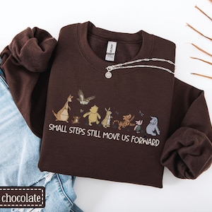 May include: A dark chocolate-coloured sweatshirt featuring a cartoon character graphic and the text "SMALL STEPS STILL MOVE US FORWARD." A silver necklace with a circular pendant rests on the sweatshirt. The sweatshirt is styled with blue jeans.