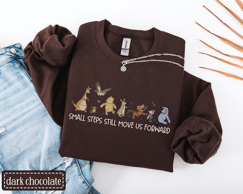 May include: A dark chocolate-coloured sweatshirt featuring a cartoon character graphic and the text "SMALL STEPS STILL MOVE US FORWARD." A silver necklace with a circular pendant rests on the sweatshirt. The sweatshirt is styled with blue jeans.