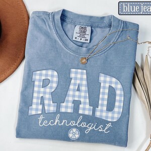 May include: Blue jean colored t-shirt with the word "RAD" in large, light blue gingham letters. The word "technologist" is printed below in white script. A gold necklace and a dried plant are also visible.