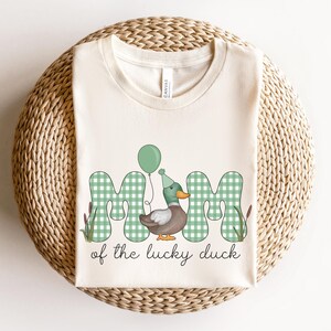 May include: A white t-shirt with a green, white, and brown graphic of a duck wearing a party hat and holding a balloon. The graphic says "Mom of the lucky duck".