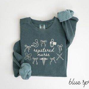 May include: Blue spruce colored sweatshirt with white graphic text and medical symbols. The text reads "registered nurse" surrounded by illustrations of a stethoscope, bandages, and syringes. The sleeves are rolled up.