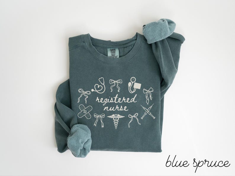 May include: Blue spruce colored sweatshirt with white graphic text and medical symbols. The text reads "registered nurse" surrounded by illustrations of a stethoscope, bandages, and syringes. The sleeves are rolled up.