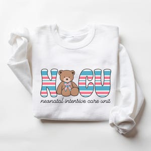 Puede incluir: Camiseta blanca de manga larga con un gr&aacute;fico de un osito de peluche en el centro del texto "NICU" con rayas azules y rosas. El texto "unidad de cuidados intensivos neonatales" est&aacute; debajo del gr&aacute;fico.