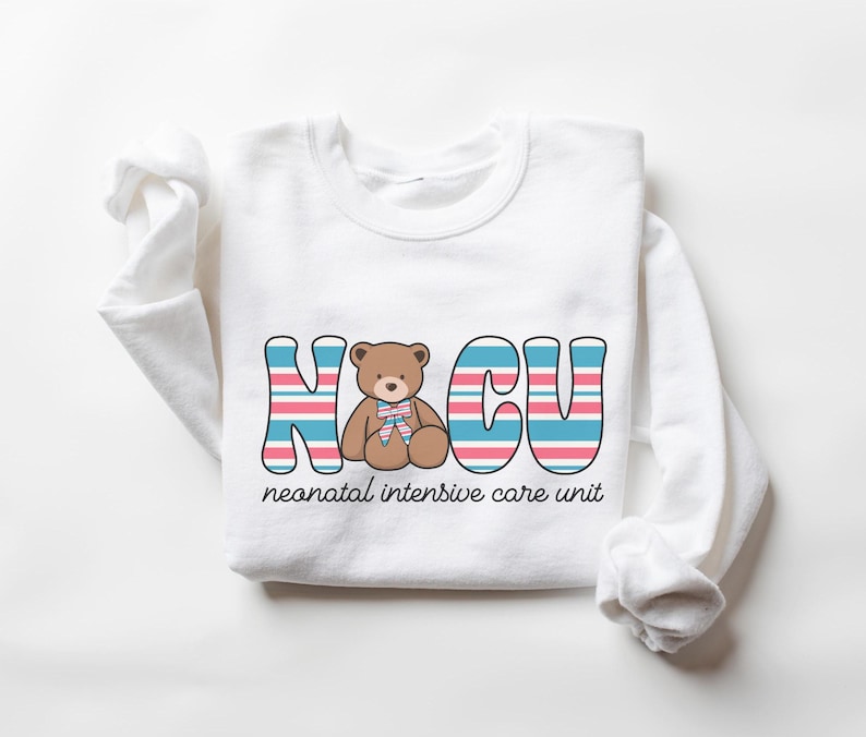 Puede incluir: Camiseta blanca de manga larga con un gr&aacute;fico de un osito de peluche en el centro del texto "NICU" con rayas azules y rosas. El texto "unidad de cuidados intensivos neonatales" est&aacute; debajo del gr&aacute;fico.