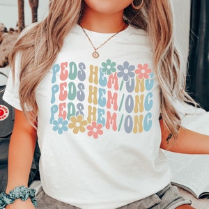 May include: A white t-shirt with the text "PEDS HEM/ONG" repeated three times in a retro font. The text is in different colors, including pink, blue, yellow, and green. Each line of text is decorated with a colorful flower.
