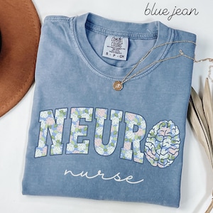 May include: A blue jean Comfort Colors t-shirt with the word "NEUR" in a floral pattern and a brain graphic. The word "nurse" is written in cursive below the word "NEUR". A gold necklace with a pendant is on the shirt.