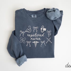 May include: A denim blue sweatshirt with white text and illustrations. The words "registered nurse" are surrounded by medical symbols, including a stethoscope, band-aid, and syringe. The word "denim" is written in cursive in the bottom right corner.