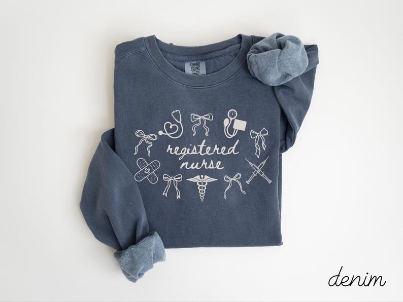 May include: A denim blue sweatshirt with white text and illustrations. The words "registered nurse" are surrounded by medical symbols, including a stethoscope, band-aid, and syringe. The word "denim" is written in cursive in the bottom right corner.