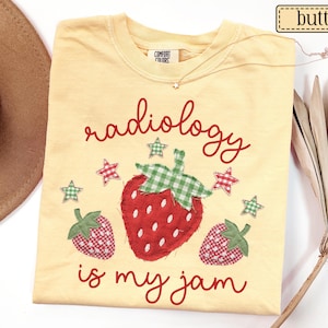 May include: A butter yellow t-shirt featuring the red text "radiology is my jam" and three strawberry graphics with green and red gingham fabric patterns. The design includes small green and red gingham stars surrounding the strawberries.