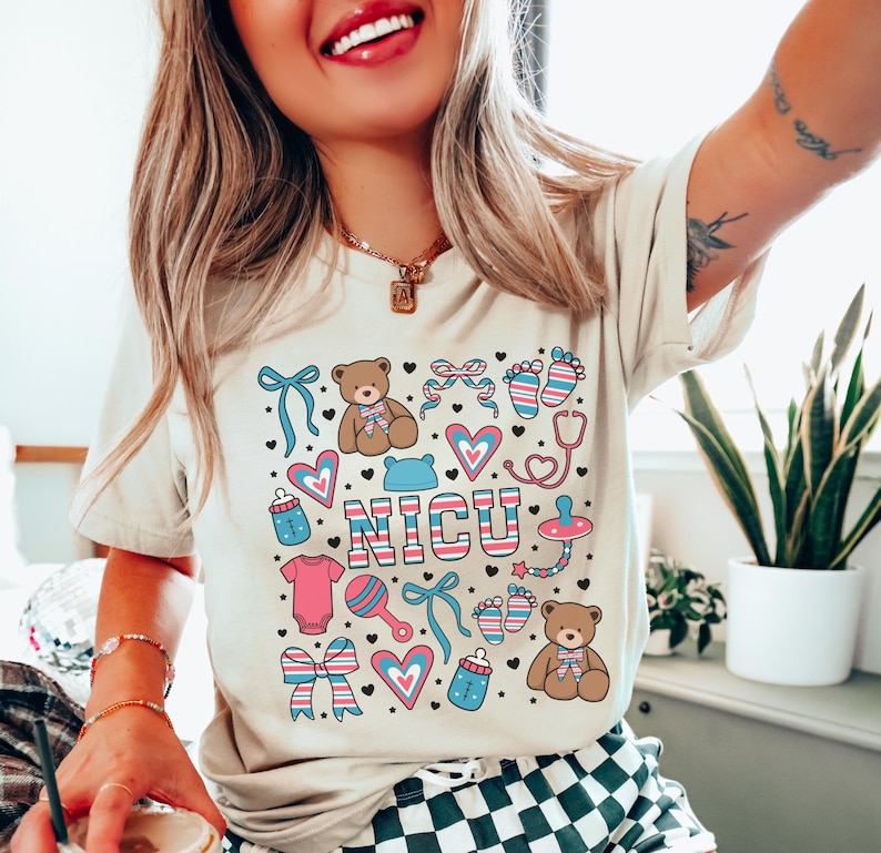 May include: A light beige t-shirt with a graphic design of a pink and blue baby footprint, a stethoscope, a teddy bear, a rattle, a pacifier, a bottle, a bow, and the word "NICU" in pink and blue letters.