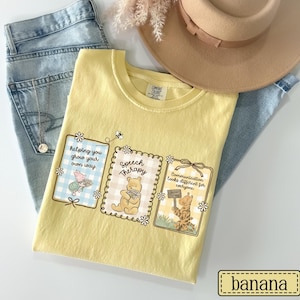 May include: A yellow Comfort Colors t-shirt featuring three framed illustrations with Winnie the Pooh characters and the phrases "Helping you grow your own way," "Speech Therapy," and "Communication looks different for everyone." The shirt is paired with light wash jeans and a tan hat.