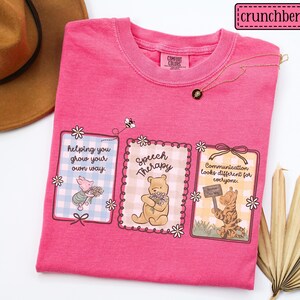 May include: A hot pink t-shirt with three framed illustrations featuring cartoon characters and floral accents. The illustrations include text that reads "helping you grow your own way," "Speech Therapy," and "Communication looks different for everyone."