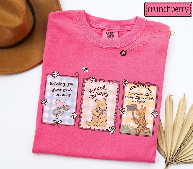 May include: A hot pink t-shirt with three framed illustrations featuring cartoon characters and floral accents. The illustrations include text that reads "helping you grow your own way," "Speech Therapy," and "Communication looks different for everyone."