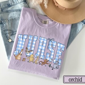 K&ouml;nnte beinhalten: Orchideenfarbenes T-Shirt mit dem Wort "NURSE" in blau-wei&szlig; karierten Buchstaben. Das Shirt zeigt Zeichentrickfiguren unter dem Text. Ein beiger Hut und blaue Jeans sind im Hintergrund.
