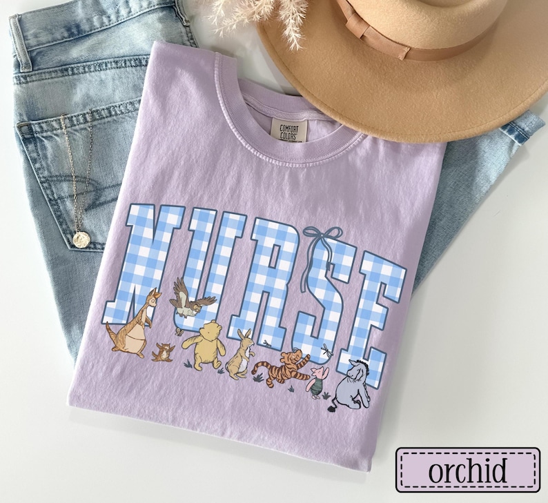 K&ouml;nnte beinhalten: Orchideenfarbenes T-Shirt mit dem Wort "NURSE" in blau-wei&szlig; karierten Buchstaben. Das Shirt zeigt Zeichentrickfiguren unter dem Text. Ein beiger Hut und blaue Jeans sind im Hintergrund.