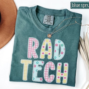 May include: A teal Comfort Colors t-shirt with the words "RAD TECH" in a colorful, patchwork design. The letters are outlined with a scalloped edge and decorated with floral and gingham patterns. A gold necklace with a pendant rests on the shirt.