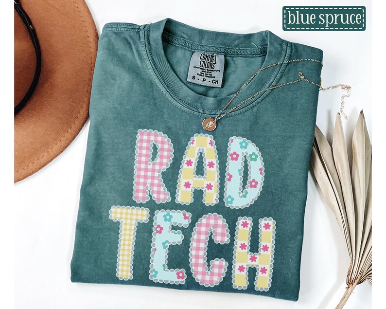 May include: A teal Comfort Colors t-shirt with the words "RAD TECH" in a colorful, patchwork design. The letters are outlined with a scalloped edge and decorated with floral and gingham patterns. A gold necklace with a pendant rests on the shirt.