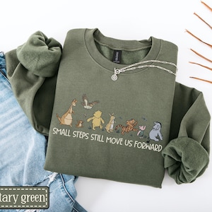 May include: Military green sweatshirt with a crew neck and long sleeves. The sweatshirt features an illustration of cartoon characters and the text "SMALL STEPS STILL MOVE US FORWARD". A silver necklace is draped on the sweatshirt.
