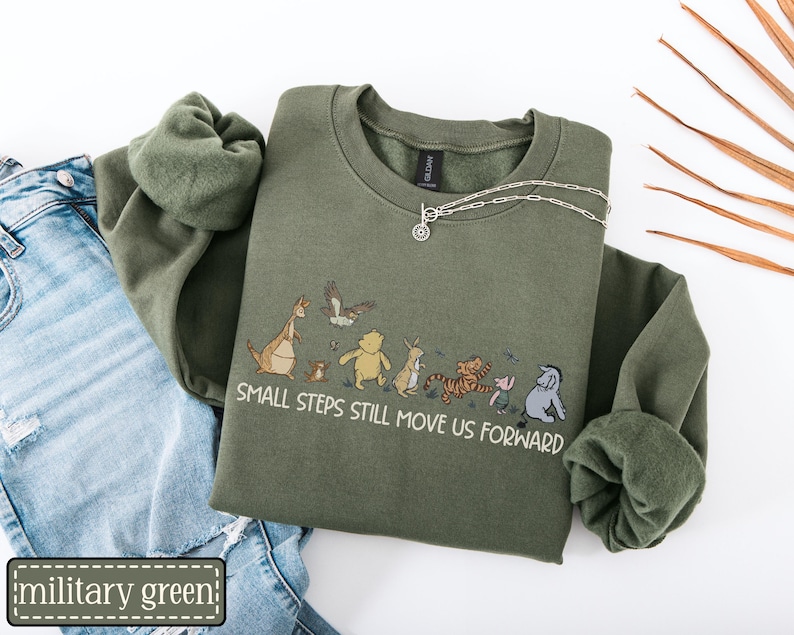 May include: Military green sweatshirt with a crew neck and long sleeves. The sweatshirt features an illustration of cartoon characters and the text "SMALL STEPS STILL MOVE US FORWARD". A silver necklace is draped on the sweatshirt.