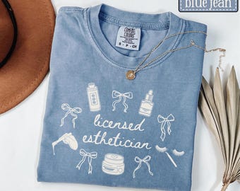 May include: A blue short-sleeve t-shirt featuring white line art graphics of bows, skincare bottles, a jar, and eyelashes. The text "licensed esthetician" is printed in the center. A gold-toned necklace with a small circular pendant rests on the collar.
