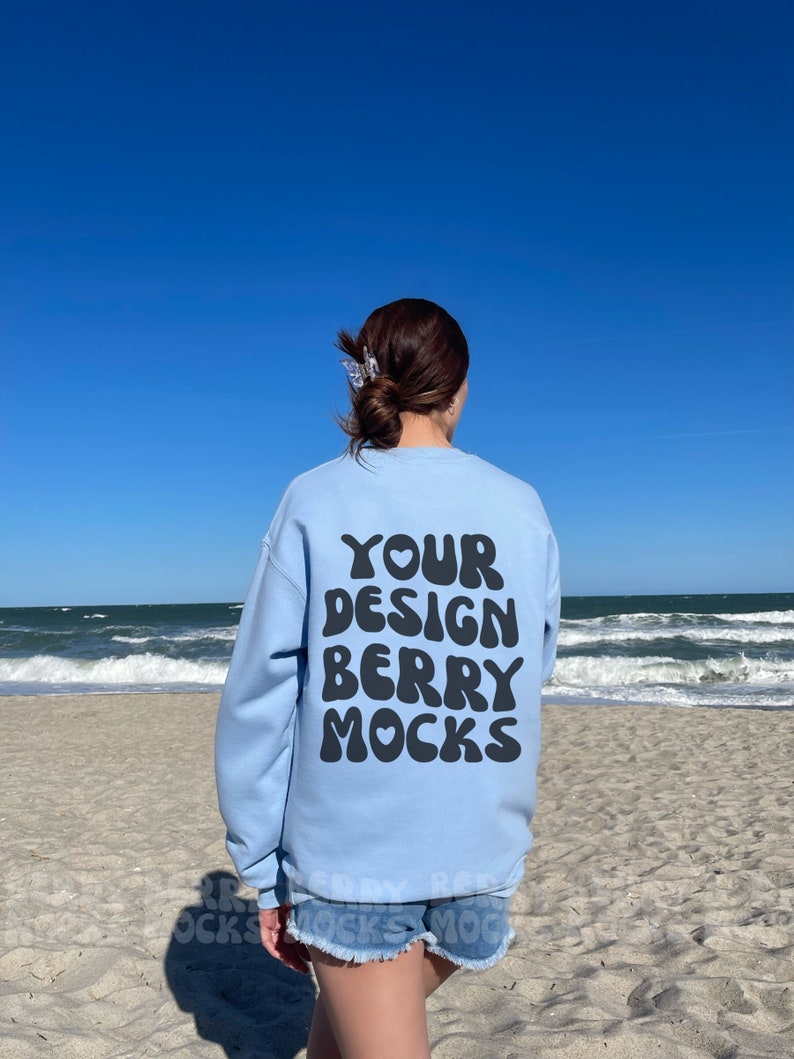 May include: A light blue sweatshirt with the text "YOUR DESIGN BERRY MOCKS" in black bubble letters on the back. The sweatshirt is being worn by a person on a beach.
