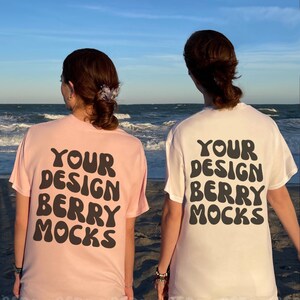 Może przedstawiać: Dwie osoby w różowych i białych koszulkach z napisem "Your Design Berry Mocks" na plecach. Osoby stoją na plaży z oceanem w tle.