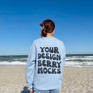 May include: A light blue sweatshirt with the text "YOUR DESIGN BERRY MOCKS" printed on the back. The sweatshirt is being worn by a person on a beach.