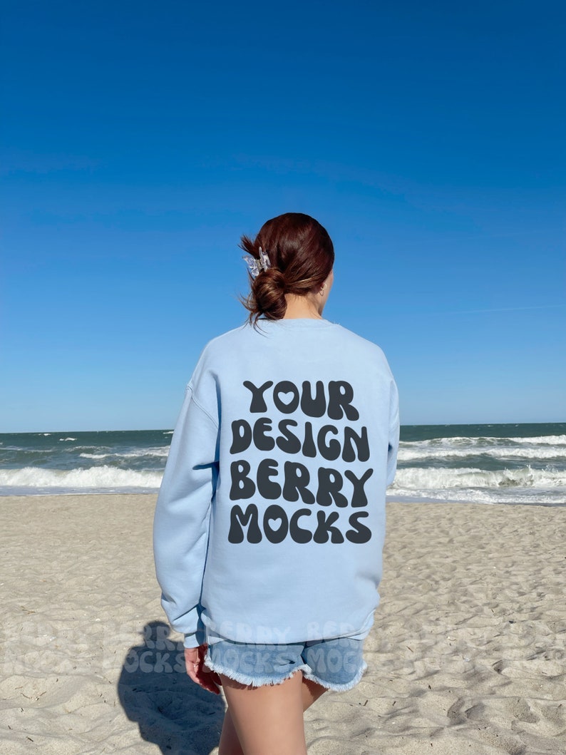 May include: A light blue sweatshirt with the text "YOUR DESIGN BERRY MOCKS" printed on the back. The sweatshirt is being worn by a person on a beach.