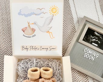 Puede incluir: Una caja de regalo blanca que contiene dos calcetines de bebé de felpa beige con caras de oso, sobre papel triturado. La tapa muestra una ilustración de una cigüeña, un sol, nubes y el texto: November 2025, Baby Philip's Coming Soon! Al lado, un tablero.