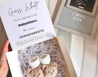Puede incluir: Una caja de regalo blanca que contiene dos patucos de bebé con forma de osito beige y puños acanalados blancos, sobre papel triturado blanco. La tapa de la caja muestra el texto: Guess What? You're going to be Babysitters We mean... GRANDPARENTS!