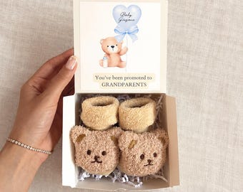 Puede incluir: Una caja de regalo blanca que contiene dos calcetines de bebé de color tostado con caras y orejas de oso. La tapa interior presenta un gráfico de un osito de peluche con un globo azul, el texto Baby Gingane y la frase You've been promoted to GRANDPARENTS.