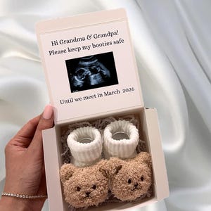 May include: A gift box containing baby booties. The box is open, revealing two pairs of white and brown teddy bear-shaped baby socks. The box lid has text that reads "Hi Grandma & Grandpa! Please keep my booties safe Until we meet in March 2026."